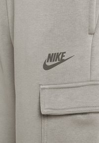 Šedé tepláky vyrobené z měkkého materiálu, s boční kapsou a černým logem Nike vytištěným v oblasti stehna.