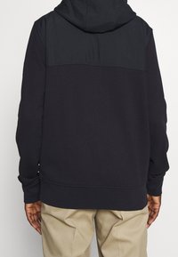 Sudadera negra con una sección superior en contraste, puños acanalados y un bolsillo frontal tipo canguro; confeccionada con una tela suave y texturizada, con un ajuste relajado.