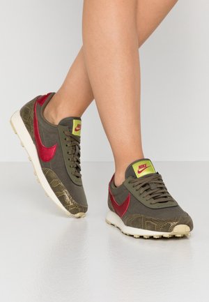 Personne portant des baskets Nike vert olive avec un swoosh rouge et un logo jaune-vert sur une semelle blanche, debout sur un sol blanc.