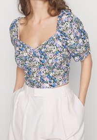 Blommig crop top i pastellfärger med puffärmar, prydd med en blandning av blå, rosa och gröna blommor mot en ljus bakgrund. Strukturerad tyg.