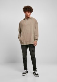 Béžový pulóver s kapucňou a polovičným zipsom vpredu, s prednými vreckami a rebrovanými rukávmi, v kombinácii s tmavými, worn-out skinny džínsami a čiernymi teniskami.