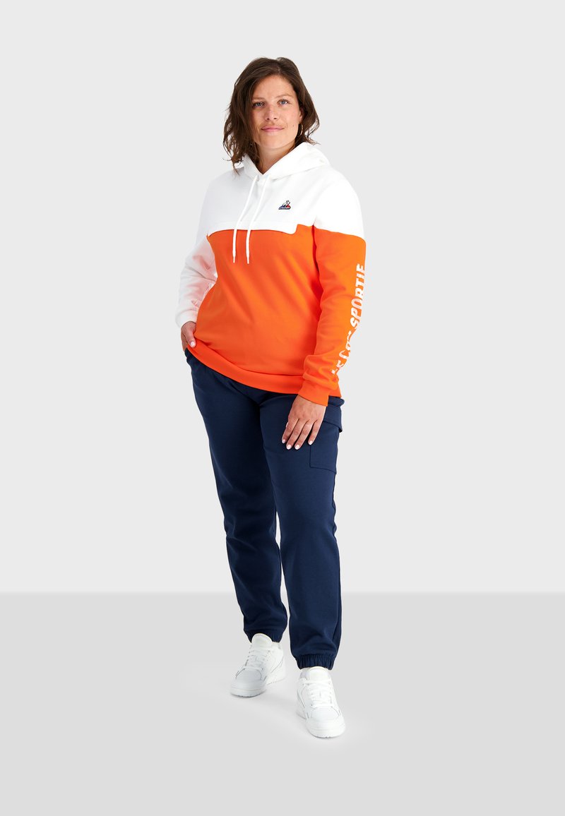 Sweat-shirt à capuche orange et blanc avec des cordons, arborant un logo et un texte sur la manche. Assorti à des joggings bleu marine et des baskets blanches.