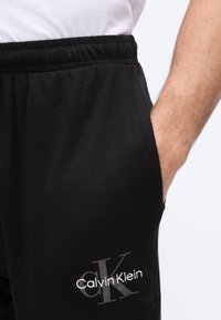 Άνδρας που φοράει μαύρο παντελόνι φόρμας Calvin Klein με ελαστική μέση και χέρι στην πλαϊνή τσέπη, συνδυασμένο με λευκό πουκάμισο.