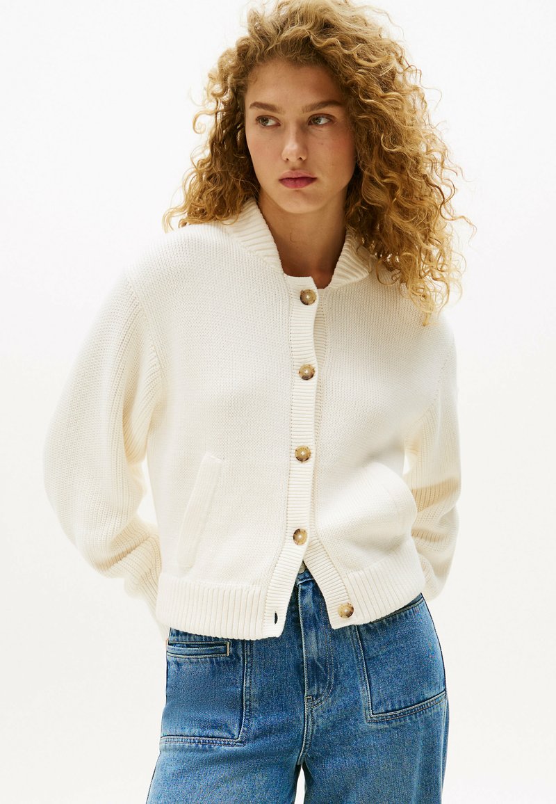 White knitted cardigan with a ribbed collar and cuffs, featuring large buttons and two front pockets. Paired with blue denim jeans.