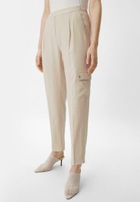 Beżowe spodnie o dopasowanym kroju z plisowanym przodem, kieszeniami bocznymi i luźnym fasonem. W zestawieniu z plecionymi, szpiczastymi mule.