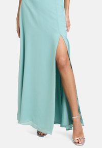 Długa, miętowa sukienka z lekkiego materiału z wysokim rozcięciem z boku, odsłaniającym nogę, zestawiona z metalicznymi srebrnymi sandałami.