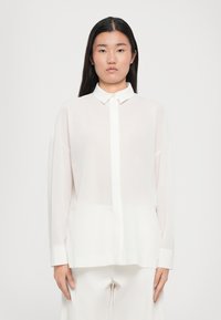 Hvit semi-transparente bluse med spiss krage, lange ermer og knappefront. Glatt tekstur med løs, avslappet passform.
