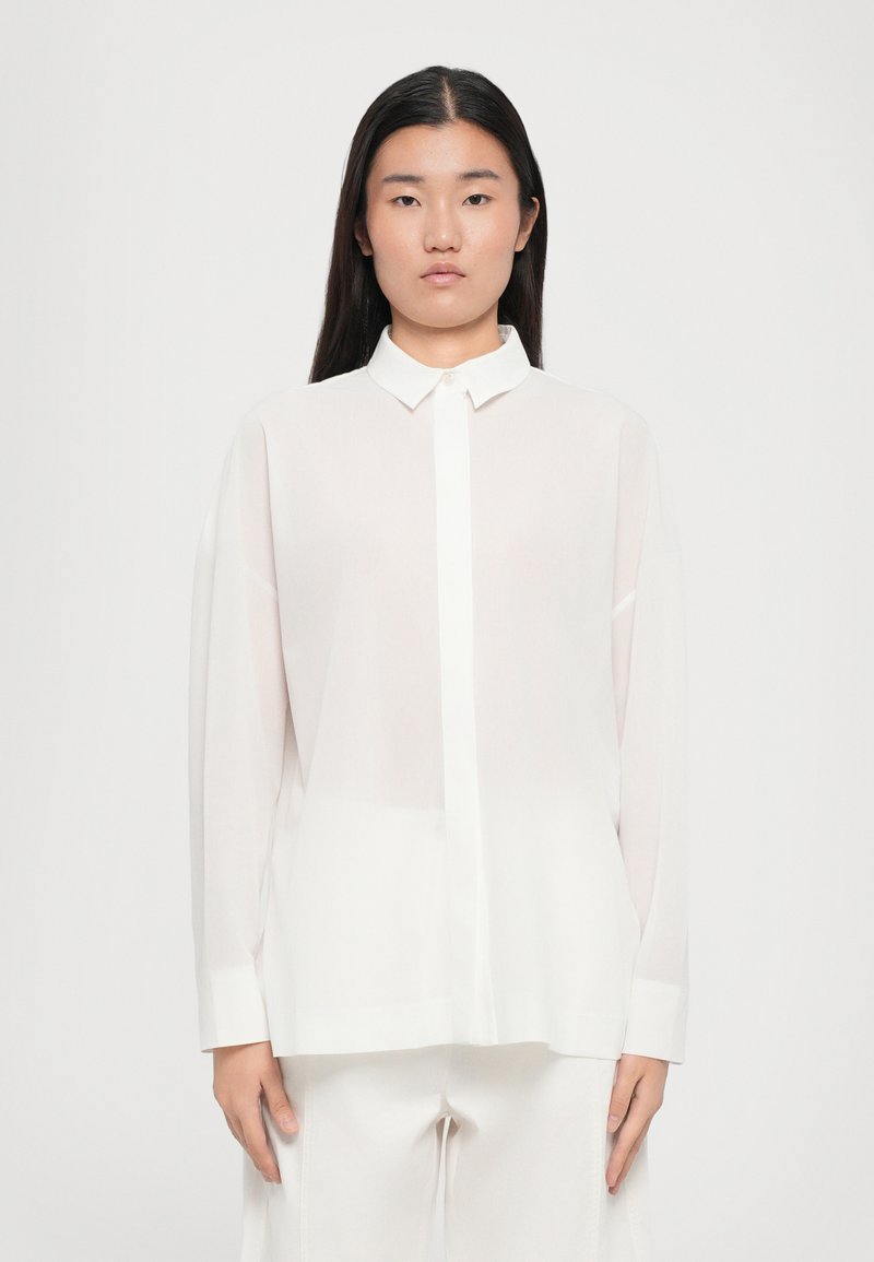 Hvit semi-transparente bluse med spiss krage, lange ermer og knappefront. Glatt tekstur med løs, avslappet passform.