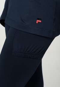 Navy sininen aktiivinen vaatetussetti, joka sisältää tekstuurilla varustetun topin matalalla helmalla ja tiukat legginsit, joissa on ryppy detail nilkassa. Punainen ja musta logo korostus.