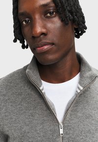 Szary sweter zapinany na zamek z wysokim kołnierzem, o miękkiej fakturze, z srebrnym zamkiem błyskawicznym, w zestawieniu z białą koszulką pod spodem.