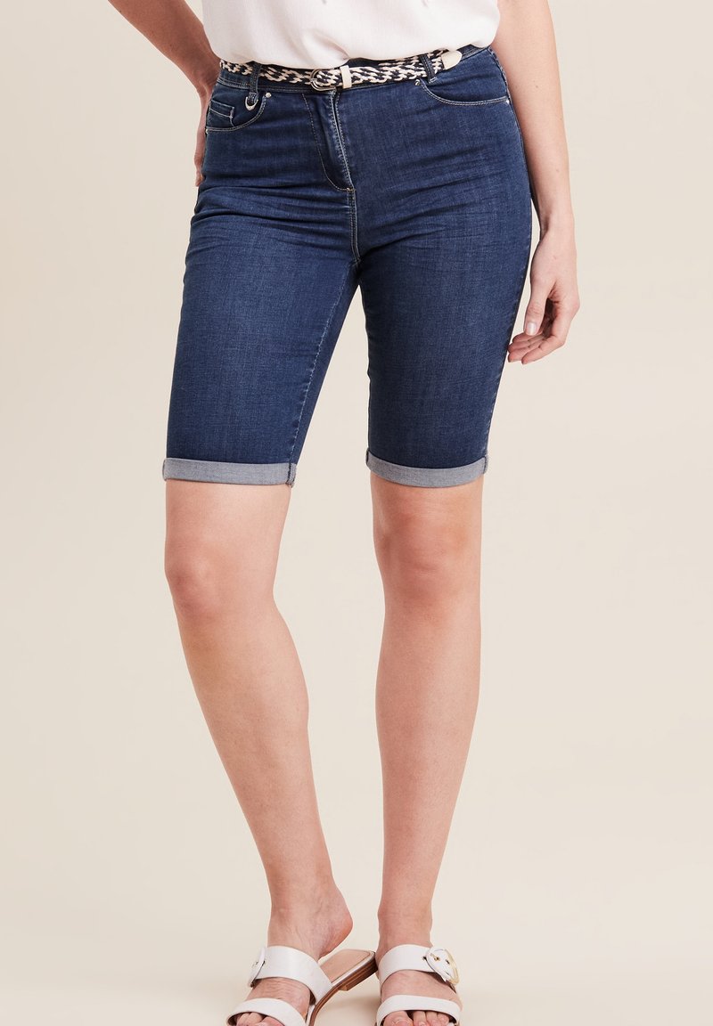 Donkerblauwe denim Bermuda shorts met omgeslagen zoom, vijf zakken en subtiele stiksels, gecombineerd met lichtgekleurde sandalen.