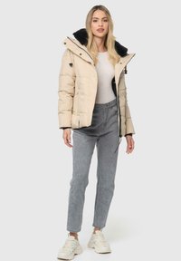 Beige puffer jacket with a detachable black fur-lined hood, zip fastening, and quilted pattern. Paired with gray high-waisted jeans and sneakers.