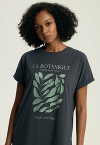 Ciemnoszara koszulka z grafika zielonego liścia oraz napisem "LA BOTANIQUE Galerie de la Nature" i "Beaute des Plantes" w kolorze kremowym. Krótkie rękawy.