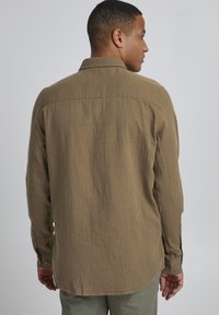 Chemise marron à manches longues en tissu texturé avec un col à rabat, dotée de boutons aux poignets et d'un ourlet arrondi, vue de dos.