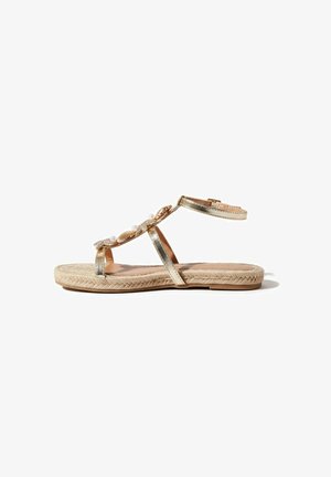 Sandalia plana con tiras metálicas doradas, hebilla en el tobillo, decorada con perlas y conchas, y suela tejida en beige.