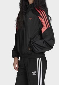 Svart träningsjacka med rosa och svarta ränder, dragkedja, ståkrage och en Adidas-logga på bröstet. Matchande svarta träningsbyxor med vita ränder.