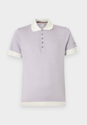 Jeune homme debout, mains dans le dos, portant un polo violet clair, un pantalon beige et des baskets blanches sur un fond blanc uni.