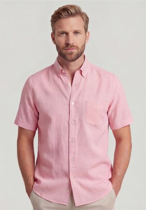 Man met kort blond haar en baard, gekleed in een lichtroze overhemd met korte mouwen en een borstzak, staand met handen in de zakken.