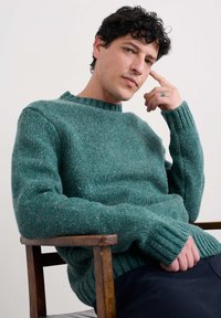 Tirkizni pleteni pulover s okruglim vratom, rebrastim manžetama i teksturiranim izgledom, u kombinaciji s tamnim hlačama, sjedi na drvenoj stolici.