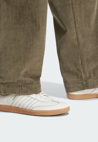 White sneaker with grey stripes and gum sole, paired with wide, olive green corduroy pants featuring a ribbed texture and cuffed hems.