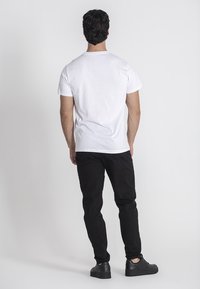 Muškarac stoji okrenut leđima, obučen u jednostavnu bijelu majicu, crne hlače i crne tenisice na jednostavnoj bijeloj pozadini.