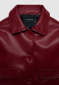 Veste en cuir bordeaux avec col, dotée de deux poches avant et de boutons rouges. Texture lisse et design élégant. Étiquette de marque "CALLIOPE" à l'intérieur.