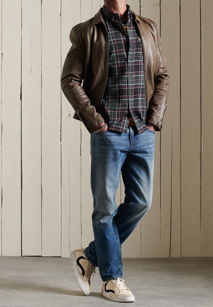 Homme portant une veste en cuir marron par-dessus une chemise à carreaux noirs et verts, jean bleu et baskets blanches, debout contre un mur en panneaux de bois.