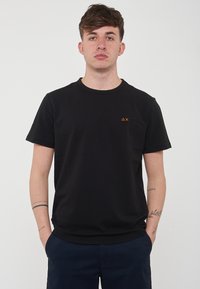T-shirt nera in cotone con collo rotondo, maniche corte e un piccolo logo arancione sul lato sinistro del petto. Texture morbida, vestibilità regolare.