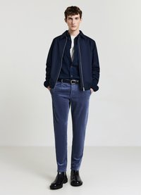 Giacca bomber navy sopra una camicia a bottoni scura, abbinata a pantaloni in velluto a coste blu e stivali neri alla caviglia. Design minimalista e snello.