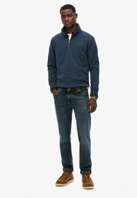 Granatowy polar z półkolem, sparowany z koszulą w kratę, niebieskimi dżinsami i brązowymi, sznurowanymi butami. Prosty design z delikatnym logo.