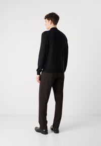 Schwarzer Pullover mit geripptem Saum, kombiniert mit dunkelbraunen Hosen. Die Textur ist glatt, und das Design umfasst einen hohen Kragen und lange Ärmel.