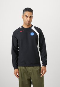 Sudadera negra de manga larga, con un panel diagonal blanco y el logo de Nike en rojo. Logo azul de París en el pecho, tejido texturizado.