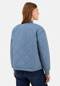 Femme aux cheveux châtain clair portant une veste matelassée bleue et un jean foncé, vue de dos sur un fond blanc.