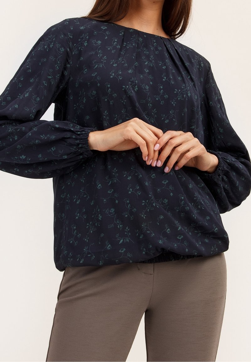 Femme portant un chemisier bleu marine à fleurs avec de longues manches bouffantes et un pantalon beige ajusté, mains délicatement jointes au niveau de la taille.