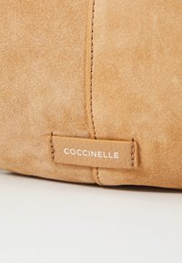 Bolso de ante en color marrón claro con costura, etiqueta de cuero rectangular con el logo "Coccinelle" en blanco en relieve. Textura suave y diseño minimalista.