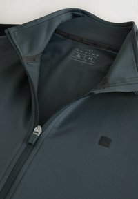 Veste zippée gris foncé avec un petit logo triangulaire sur la poitrine et la marque « Next Active Aim » à l'intérieur du col.