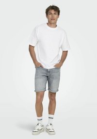 Camiseta blanca de manga corta, pantalones cortos de mezclilla azul claro con dobladillos enrollados, zapatillas blancas con detalles en gris y calcetines a rayas.
