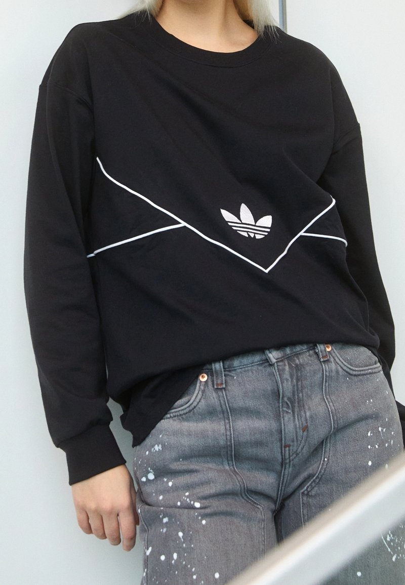 Person wearing a black Adidas sweatshirt with white piping and gray jeans with white paint splatters, standing against a pale background.