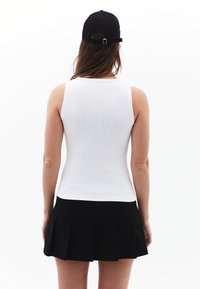 Top sin mangas blanco con textura de canalé combinado con una falda corta plisada negra. Se lleva una gorra negra puesta al revés, con detalle de correa ajustable en la parte posterior.