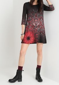 Czarna sukienka z czerwonymi kwiatowymi i paisley wzorami, z rękawami do połowy ramienia i rozkloszowanym krojem, w zestawieniu z czarnymi, sznurowanymi botkami i bordowymi skarpetkami.