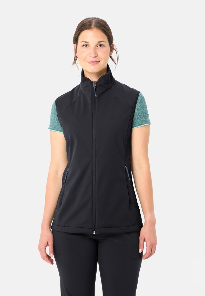 Frau trägt eine schwarze, ärmellose Weste mit Reißverschluss über einem grünen Kurzarmshirt und steht vor einem einfarbigen weißen Hintergrund.