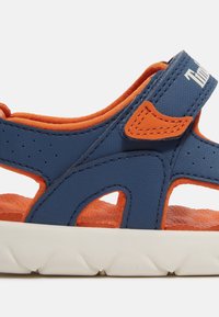 Siniset synteettiset sandaalit oransseilla yksityiskohdilla, rei'itetty päällinen, säädettävä hihna, teksturoitu pohja, ja valkoinen pehmustettu pohja.