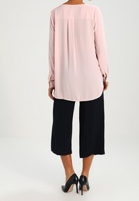 Blusa rosa chiaro con maniche lunghe, scollo rotondo e piega sul retro. Abbinata a pantaloni culotte neri a gamba larga e tacchi alti neri.