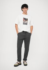 Een jonge man staat met zijn handen in zijn zakken, gekleed in een wit T-shirt met een afbeelding van een hond, donkere grijze broek en loafers.
