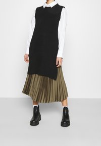 Svart stickad ärmlös tröja över en vit skjortkrage, tillsammans med en plisserad olivgrön kjol och svarta ankelstövlar.