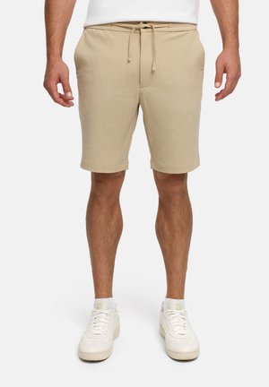 Homme portant un short beige à cordon, des baskets blanches et une chemise blanche, mains détendues le long du corps.