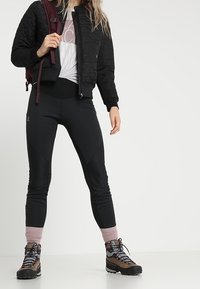 Svart vadderad jacka, figurnära svarta byxor och bruna vandringskängor med rosa detaljer. Modellen bär en vinröd ryggsäck och har på sig en vit t-shirt med grafik.