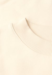 Sweat-shirt beige clair avec un col rond côtelé et une texture de tissu lisse. Aucun motif ni élément visible. Vue rapprochée.