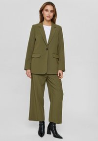 Oliivroheline bleiser kahe eesmise taskuga, ühe nupu sulgemisega, koos vabalt voolavate oliivroheliste pükste ja mustade kontsaga ankle'ite kingadega.