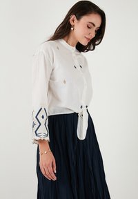 Camisa de lino blanca con mangas tres cuartos, con bordado geométrico azul y dorado en las mangas, cierre de botones y un lazo en la parte delantera.